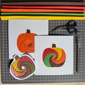 Puede incluir: Dos tarjetas cuadradas blancas con diseños de calabazas recortadas en papel con patrones en espiral, una en naranja y otra en segmentos multicolores. También se ven una pegatina de calabaza, tijeras, un cúter y tiras de papel de colores.
