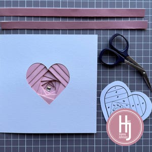 Könnte beinhalten: Hellblaue Karte mit herzförmigem Ausschnitt, der ein geschichtetes rosa Papierdesign zeigt. Zwei rosa Papierstreifen und eine Schere liegen neben der Karte. Eine herzförmige Schablone und ein rosa Logo mit "Hanne Johnsen" sind ebenfalls zu sehen.
