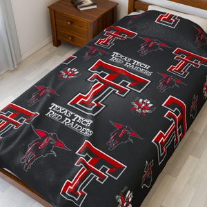 May include: Black blanket with a repeating pattern of the Texas Tech Red Raiders logo and mascot in red and white. The words "Texas Tech Red Raiders" are also visible. The blanket is on a bed.