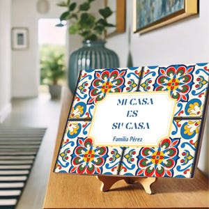 May include: A decorative tile sign with a vibrant floral pattern in red, blue, yellow, and green. The sign reads "MI CASA ES SU CASA" and "Familia Pérez" in a white frame. It is displayed on a wooden stand.