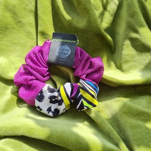 Könnte beinhalten: Ein rosa-lilafarbenes Mesh-Haargummi mit einem schwarzen und weißen Leopardenmusterstoff und einem bunten gestreiften Stoff. Das Haargummi liegt auf einer grünen Oberfläche. Das Haargummi hat ein Etikett mit der Aufschrift "Viva la Grit"