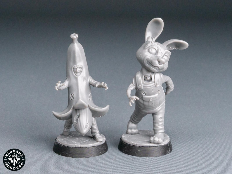 May include: Two gray miniature figurines. One is a person in a banana costume, the other is a rabbit wearing overalls. Both figures are on black circular bases. The Wargame Wonders logo is visible in the lower left corner.