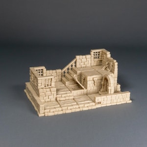 May include: A beige miniature wargame terrain piece depicting a weathered, ruined structure. The model features brickwork, archways, stairs, and window frames, suggesting a medieval or fantasy setting. The base is rectangular, and the piece is designed for tabletop gaming.