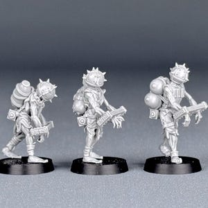 May include: Three miniature figurines of a futuristic soldier, each standing on a black base. The soldiers wear helmets with spikes, backpacks, and carry a weapon. The figures are a light gray color.