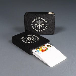Könnte beinhalten: Schwarzes Kartenetui mit dem Text "WARGAME WONDERS" in Weiß. Das Etui ist geöffnet und zeigt ein Kartenspiel mit einer bunten Jokerkarte. Dahinter befindet sich ein zweites geschlossenes Etui.