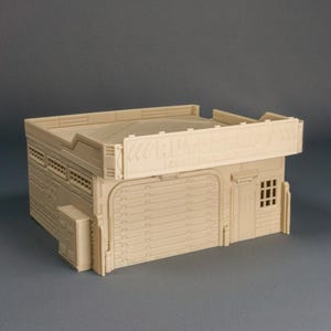 May include: A beige miniature building with a garage door, small window, and a sign that reads "Wargame Wonders". The building has a flat roof and is likely a model for tabletop gaming or a diorama.