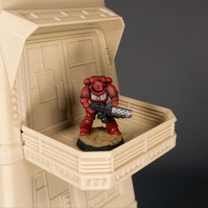 May include: A red and silver painted miniature space marine figure stands on a beige balcony with a railing. The figure is holding a gun and is standing on a small base.