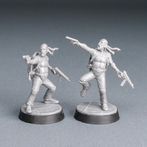 Könnte beinhalten: Zwei graue Miniaturfiguren von bewaffneten Charakteren in Actionposen. Jede Figur hält eine Pistole und ein Gewehr. Sie stehen auf runden Sockeln mit schwarzem Rand. Die Figuren sind mit taktischer Ausrüstung detailliert.