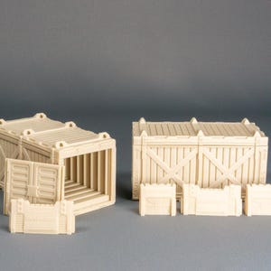 Könnte beinhalten: Beige Miniaturmodelle von Versandcontainern und Kisten. Das Set enthält zwei große Container, einer mit offenen Türen, und mehrere kleinere Kistenkomponenten. Die Modelle sind für Tabletop-Spiele oder den Modellbau konzipiert.