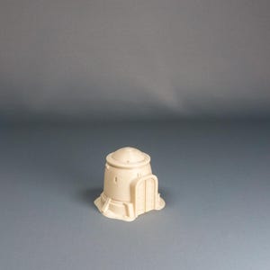 Könnte beinhalten: Ein beigefarbenes Miniaturgebäude mit einem gewölbten Dach und einem bogenförmigen Türrahmen. Das Modell hat einen zylindrischen Körper und ist wahrscheinlich ein Geländestück für Tabletop-Spiele. Der Sockel ist rechteckig.