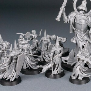 May include: A collection of unpainted miniature figures, likely for tabletop gaming. The figures are primarily gray, featuring detailed armor, flowing robes, and various weapons. Some figures have tall, pointed hats. The figures are on black bases.