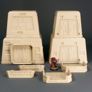 May include: A set of beige plastic building blocks for a tabletop wargame. The set includes two tall buildings with doors and windows, two short walls, and a small platform. A red plastic miniature figure stands on the platform.