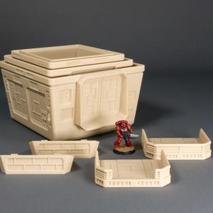 May include: A set of modular, beige plastic building pieces for tabletop gaming. The pieces include a multi-level building with windows and a flat roof, and three separate wall sections.