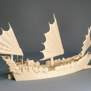 Op de afbeelding: Een gedetailleerd, crèmekleurig model van een schip met gestileerde zeilen en sierlijke details. Het schip heeft meerdere masten met grote, vleugelachtige zeilen en een gedetailleerde romp. Het model staat tegen een neutrale grijze achtergrond.