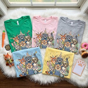 May include: Five colorful t-shirts in green, pink, gray, blue, and yellow, each featuring a cartoon group of animal characters. The shirts are arranged on a white, fluffy surface, with a carrot toy and donuts nearby. The shirts are likely for children.