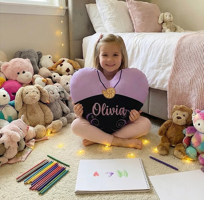 May include: A heart-shaped pillow with a black base and a lavender top, featuring the name "Olivia" in cursive. The pillow has a seashell pendant design and is surrounded by stuffed animals.