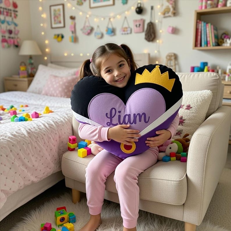 May include: A heart-shaped pillow with a black top and purple bottom, featuring a gold crown and the name "Liam" in script. The pillow has a white ribbon detail and a gold ring. The child is wearing pink pajamas.
