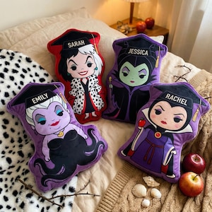 May include: Four character-shaped throw pillows. Each pillow features a different cartoon villain with a graduation cap and a name. The pillows are in shades of red, purple, and black, with details in white and gold. The pillows are on a bed with a patterned blanket.
