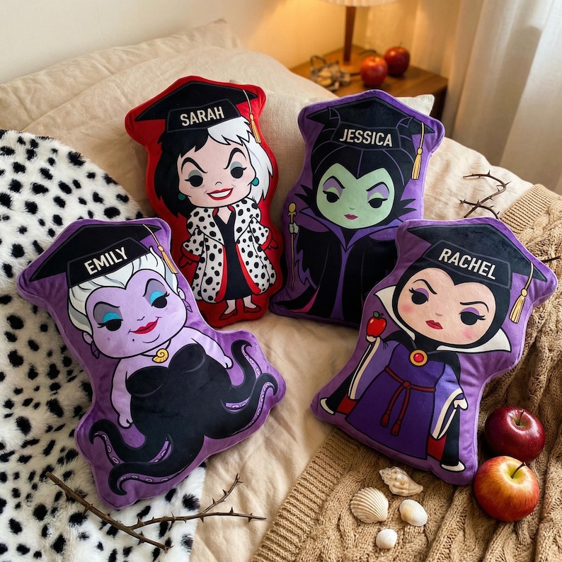 May include: Four character-shaped throw pillows. Each pillow features a different cartoon villain with a graduation cap and a name. The pillows are in shades of red, purple, and black, with details in white and gold. The pillows are on a bed with a patterned blanket.