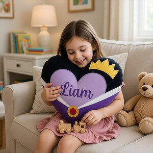 May include: A heart-shaped pillow with the name "Liam" in purple script, framed by black and purple fabric, and a gold crown. A wooden toy train sits below. The pillow is held by a child.