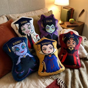 May include: Five plush cushions featuring cartoon illustrations of Disney villains. Each cushion is shaped and coloured to resemble a specific character, with names like "Emma," "Chloe," and "Audrey" printed on them. The cushions are arranged on a bed.