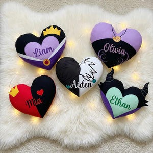 May include: Heart-shaped decorative pillows in various colors, each with a unique design and name. The pillows feature designs with crowns, horns, and other embellishments. Names include Liam, Olivia, Mia, Aiden, and Ethan.