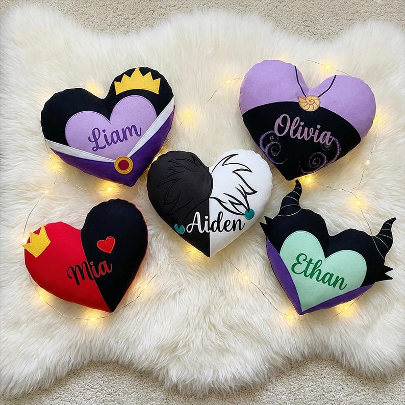 May include: Heart-shaped decorative pillows in various colors, each with a unique design and name. The pillows feature designs with crowns, horns, and other embellishments. Names include Liam, Olivia, Mia, Aiden, and Ethan.