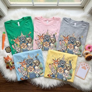 Może przedstawiać: Pięć kolorowych koszulek w kolorze zielonym, r&oacute;żowym, szarym, niebieskim i ż&oacute;łtym, każda z grupą postaci z kresk&oacute;wek. Koszulki są złożone na białej, puszystej powierzchni, z zabawką w kształcie marchewki i pączkami w pobliżu.