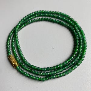 Op de afbeelding: Een ketting bestaande uit kleine, ronde, smaragdgroene kralen. De kralen zijn aan elkaar geregen en vormen meerdere strengen. Een goudkleurige sluiting zet de ketting vast. De ketting wordt gepresenteerd tegen een effen, lichtgekleurde achtergrond.