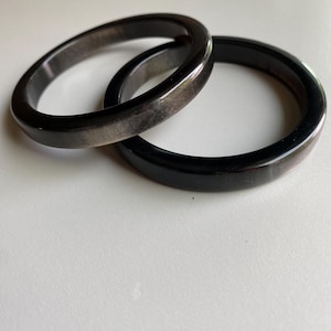 May include: Two smooth, circular bangles with a polished finish. The bracelets are dark charcoal gray and black in color, featuring subtle, natural-looking variations in tone and texture throughout the material, creating a sleek, rounded appearance.