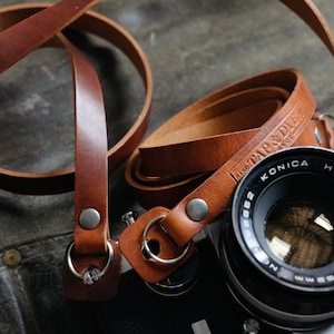 May include: A brown leather camera strap with a silver metal ring and a stamped logo that reads "Hand Tap & Die". The strap is wrapped around a black camera with a silver lens.