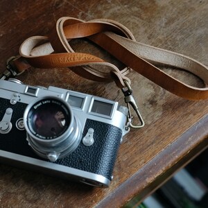 May include: A silver and black 35mm film camera with a leather strap. The camera has a lens with a silver rim and a black lens cap. The camera is sitting on a wooden surface.