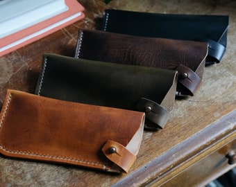 May include: Four leather eyeglass cases in different colors: brown, dark brown, green, and tan. The cases are lined up on a wooden surface.