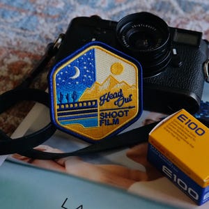 Puede incluir: Un parche bordado azul y amarillo con el texto "Head Out Shoot Film" y una escena de montaña. El parche está sujeto a una correa de cámara negra y una cámara con lente es visible en el fondo.