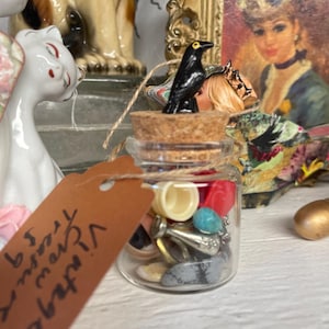 May include: A small glass jar filled with various colorful objects, including beads and buttons. The jar is topped with a cork and a small black crow figurine. A brown tag with handwritten text hangs from the jar.