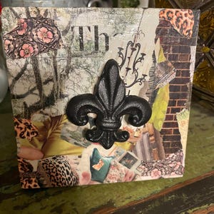 May include: A square, decorative box with a collage design. The box features a black fleur-de-lis emblem. The collage includes floral, leopard print, and brick patterns. The word "Th" is visible on the front.