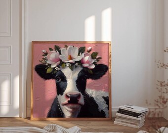 May include: A framed artwork featuring a black and white cow wearing a floral crown of pink and white flowers. The cow is set against a pink background. The artwork is in a wooden frame and is leaning against a white wall.