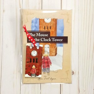 May include: A children's book titled "The Mouse and the Clock Tower" with illustrations of a mouse in a red plaid coat. The cover features a clock tower and a red and white polka dot ribbon. The book is in a clear plastic sleeve.