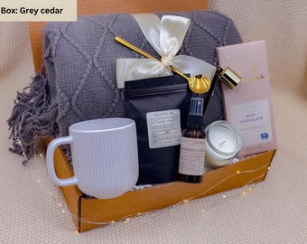 Puede incluir: Una caja de regalo con una manta gris, una taza blanca, té, chocolate, una vela y un frasco de spray. La caja está atada con una cinta blanca y una cuchara dorada. La caja está etiquetada como "Grey cedar".