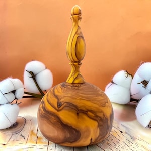 Puede incluir: Un objeto decorativo de madera con una base redondeada y lisa y una parte superior alta y delgada en forma de remate. La pieza presenta un patrón de veta distintivo y arremolinado en tonos amarillo dorado y marrón oscuro.