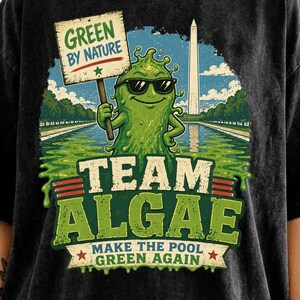 Könnte beinhalten: Ein anthrazitfarbenes, säuregewaschenes T-Shirt mit einer Grafik einer grünen Algenfigur mit Sonnenbrille, die ein Schild mit der Aufschrift GREEN BY NATURE hält. Darunter steht TEAM ALGAE und MAKE THE POOL GREEN AGAIN mit dem Washington Monument.