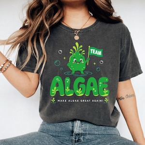 May include: A charcoal gray short-sleeve t-shirt featuring a green cartoon algae character holding a flag that says TEAM. Below the character, the word ALGAE is printed in large green letters, followed by the text MAKE ALGAE GREAT AGAIN.