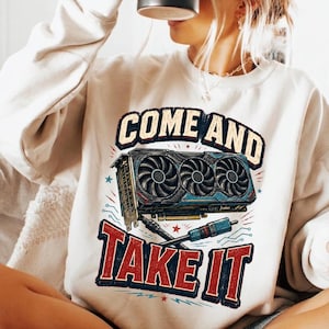 May include: A beige crewneck sweatshirt featuring a graphic print of a triple-fan graphics card positioned above a screwdriver. The text "COME AND TAKE IT" is printed in bold, distressed blue and red lettering surrounding the hardware illustration.