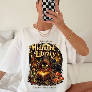 May include: A white t-shirt featuring a graphic print with the text "After Hours Midnight Library" and "Every Book Holds a Spirit." The illustration depicts a hooded figure reading a glowing book, a black raven, mushrooms, and glass bottles.