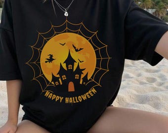 May include: Black t-shirt with a Halloween-themed graphic. The design features a full orange moon, a haunted house silhouette, bats, a witch on a broom, and the words "Happy Halloween" in a curved font. The shirt is worn by a person.