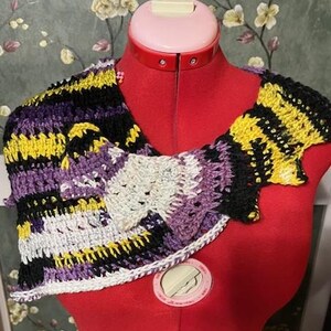 Puede incluir: Un chal de ganchillo en tonos de morado, amarillo, negro y blanco. El chal está drapeado sobre un maniquí de costura rojo.