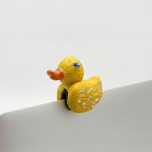 May include: A yellow duck-shaped clip with an orange beak and blue eyes, attached to a silver laptop. The duck has white speckles on its wings and is made of a textured material. The clip is designed to hold the laptop in place.