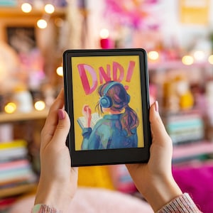 May include: A black e-reader displaying a colorful illustration with the text "DND!" on a yellow background. The illustration depicts a person wearing headphones and reading a book. The e-reader is held in front of a blurred background.