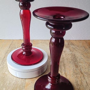 May include: Two red glass candle holders with a classic design. The candle holders have a wide base and a tall, slender stem.