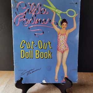 May include: Vintage "Gilda Radner Cut-Out Doll Book" on display. The cover is blue with the title in pink and yellow. A woman in a pink swimsuit holds green scissors. The book is an authorized edition and is displayed on a black stand.